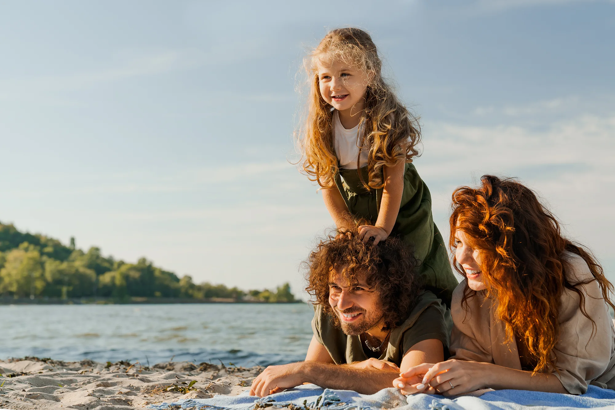 Familie am Seeufer: Ein kleines Mädchen steht lachend auf dem Rücken ihres am Strand liegenden Vaters, neben ihnen liegt eine Frau; im Vordergrund Sand und Decke, im Hintergrund Wasser und grüne Uferlandschaft im warmen Licht.