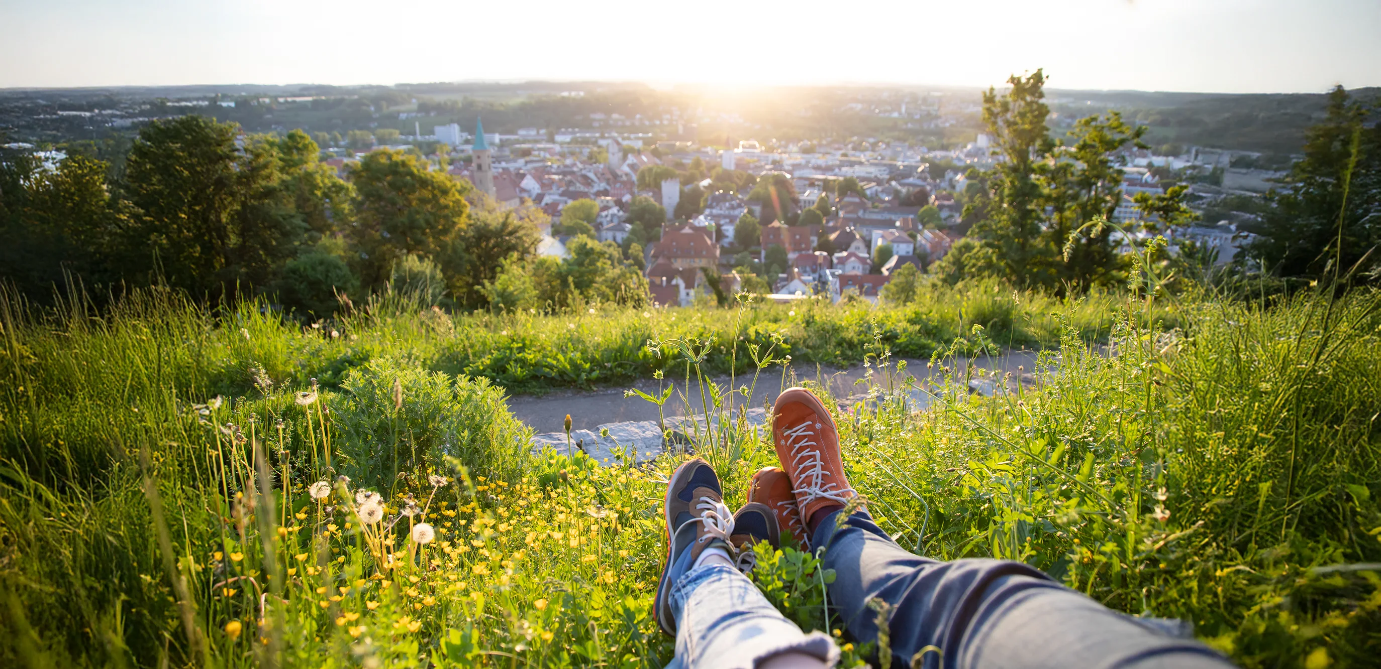 Panoramablick vom Wiesenhang auf eine Stadt im warmen Abendlicht; im Vordergrund ragen die Beine zweier Personen mit Sneakers aus dem hohen Gras und Wildblumen.