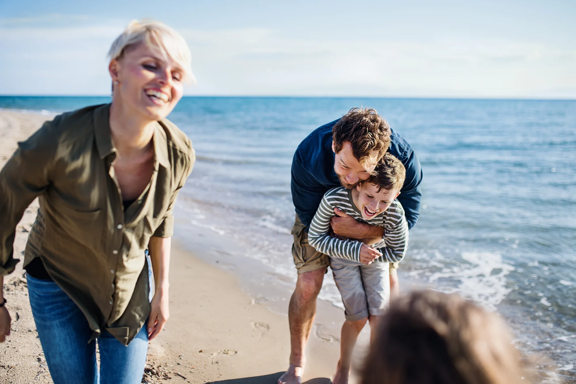 Familie am Strand: eine Frau lacht links im Bild, während ein Mann am Wasser einen Jungen von hinten umarmt und kitzelt; im Vordergrund unten unscharf eine weitere Person, dahinter Meer mit sanften Wellen.